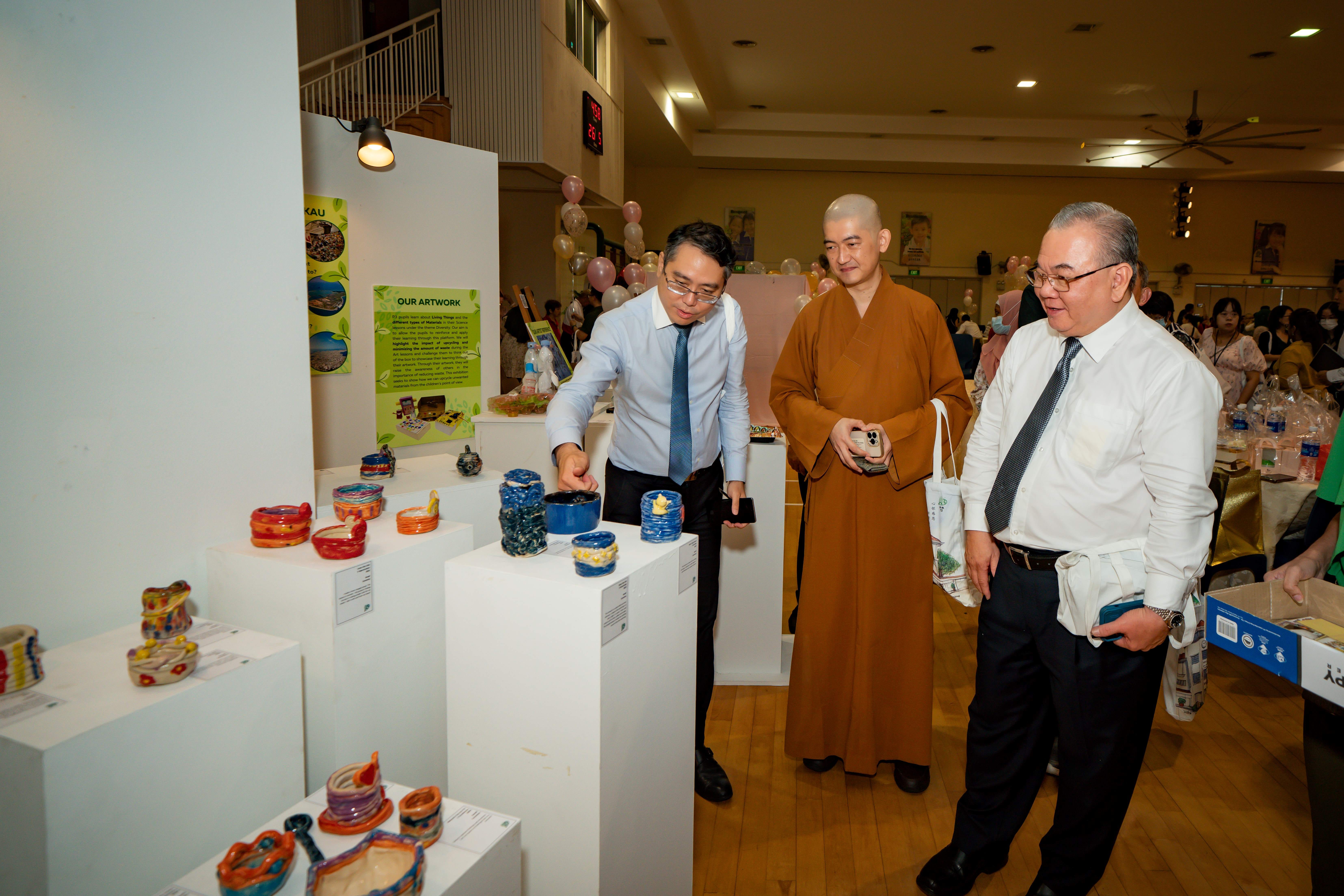 Three men, including a Buddhist monk in orange robes, examine colorful ceramic artworks displayed on white pedestals at an indoor exhibition.